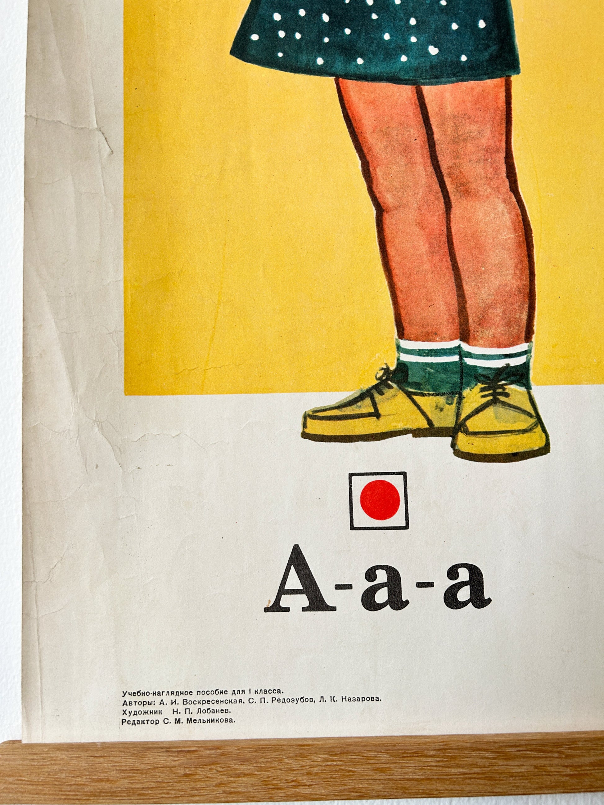 Vintage Russian educational poster showing the lowercase letter 'a' represented by the word 'A-a-a' and an illustration of a child's legs wearing yellow shoes, green socks, and a dark green polka-dot skirt against a yellow and off-white background.