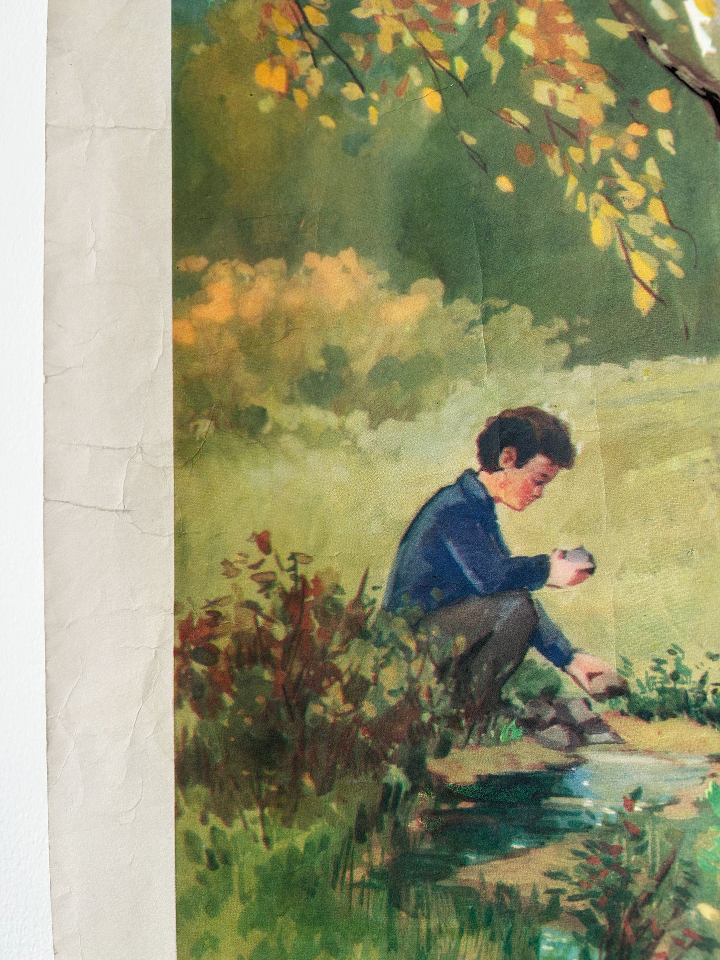 A watercolor-style art print depicting a young boy crouching by a stream in a grassy outdoor setting, with autumn leaves overhead. The print is shown with a textured off-white paper border visible on the left side.