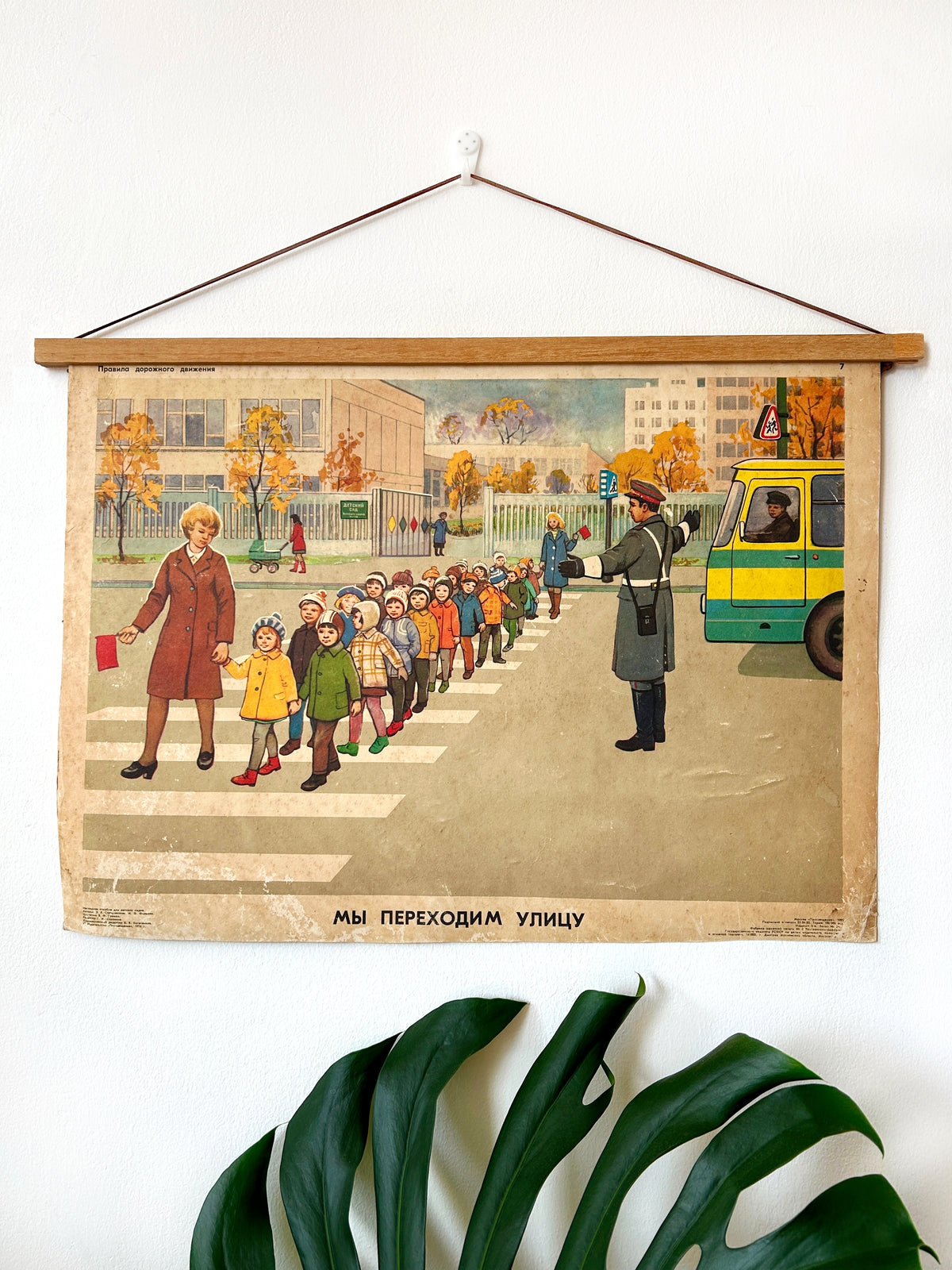 Soviet vintage educational road safety poster depicting children crossing a street with a traffic officer and a bus, suspended from a wooden rod against a white wall with a green plant leaf in the foreground.