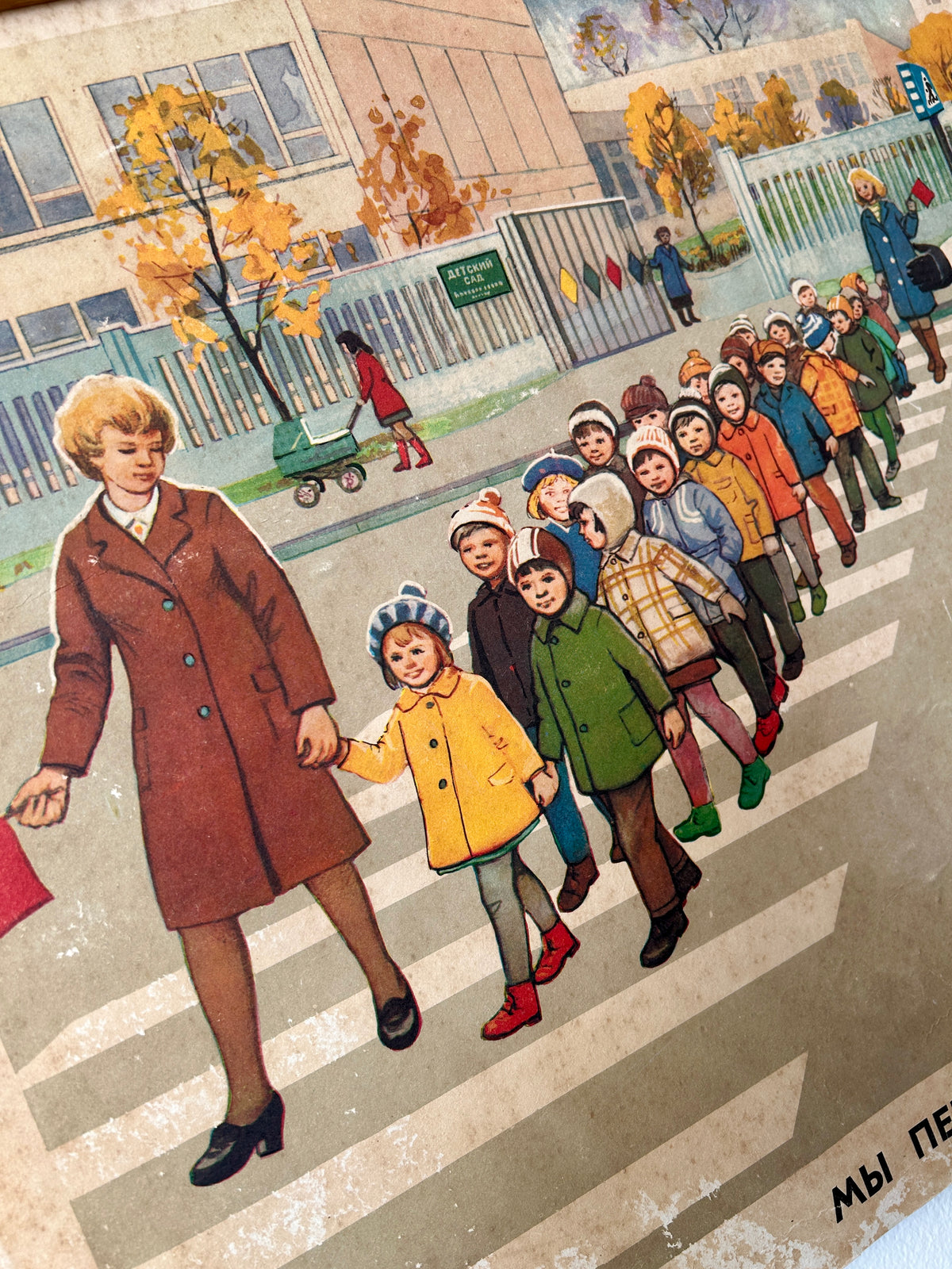 USSR vintage road safety poster detail: illustration of a woman in a brown coat holding hands with children as they cross a zebra street in front of a school building with autumn trees.