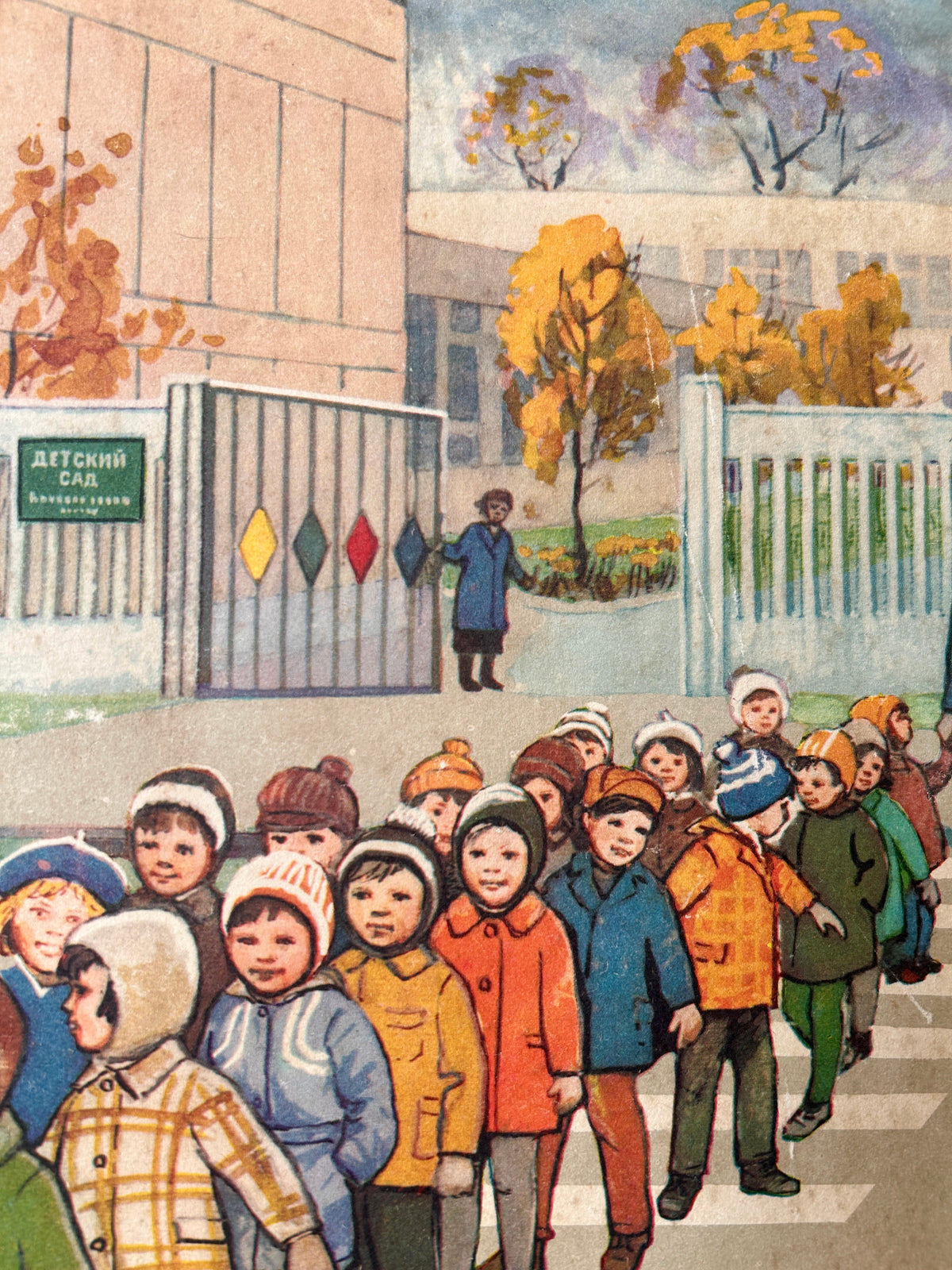 USSR vintage road safety poster detail:  illustration of a group of children in colorful winter coats walking across a crosswalk in front of a school gate and building, with autumn trees and a pale sky in the background.