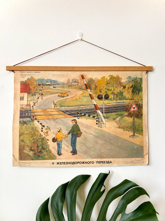 A vintage USSR railway crossing poster for children, hanging on a white wall with a wooden rod and hanging string, with green plant leaves in the foreground.