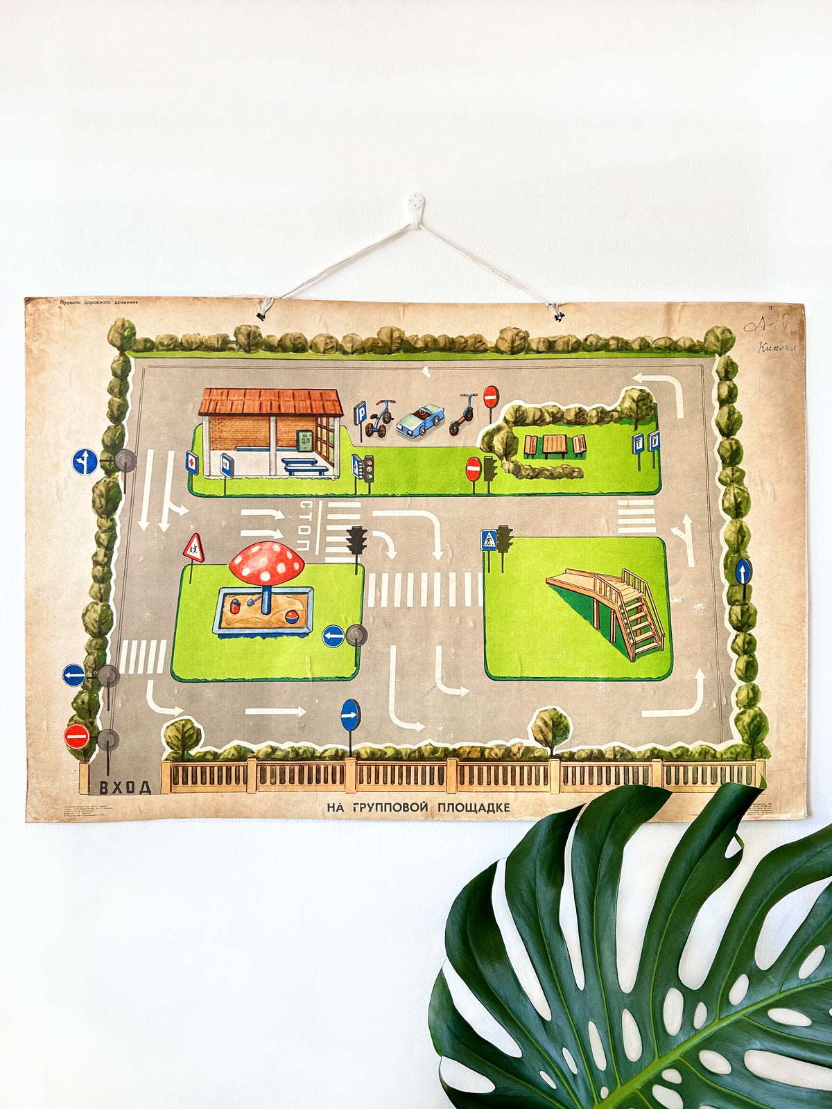 A large USSR vintage chart used in kindergartens and schools to teach children road safety, traffic signs, and pedestrian etiquette, hanging on a white wall with monsterra leaf below.