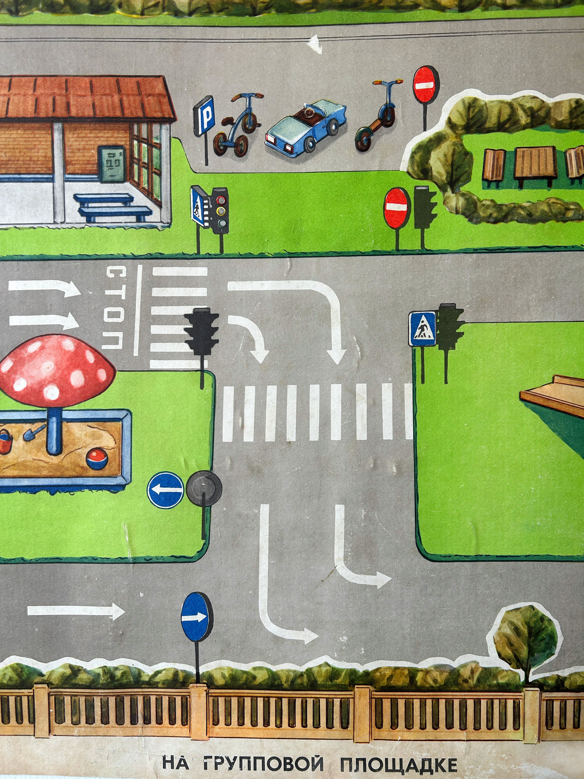 A close-up of a colourful USSR vintage children's road safety poster showing a street scene with a grey road, crosswalks, traffic lights, green grass areas, benches, a bus stop, parked scooters and a car, a mushroom-shaped sandbox, and Russian text at the bottom.