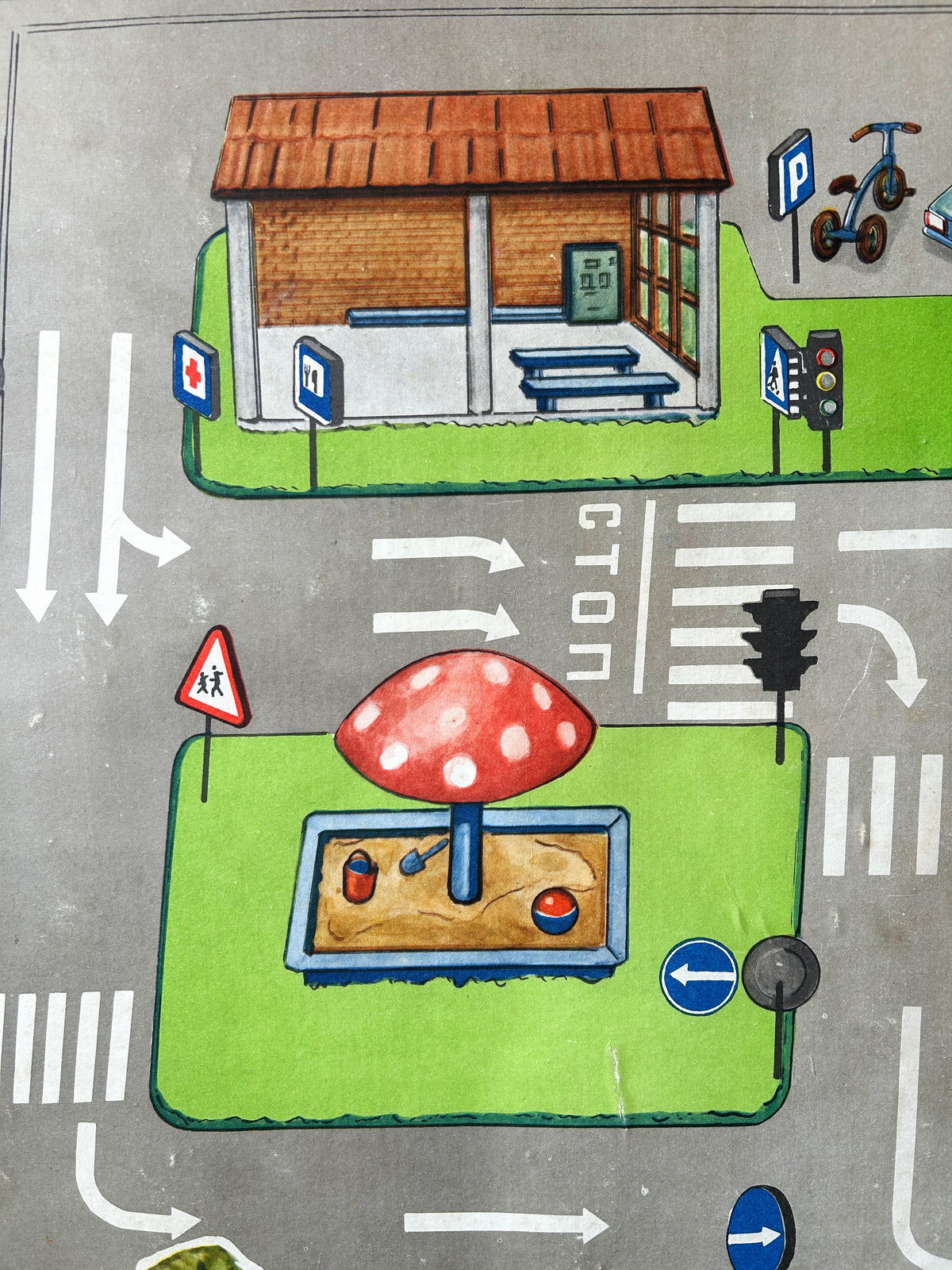A close-up of a colourful USSR vintage children's road safety poster showing a street scene with a grey road, playground with a mushroom-shaped sandbox, crosswalks, traffic lights, green grass areas, benches, a bus stop, parked scooters and a car.