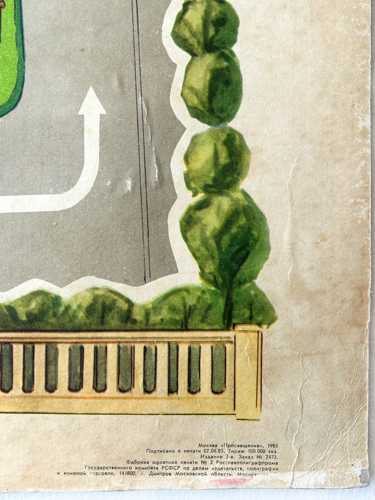 Close-up corner of a vintage USSR road safety poster showing a stylised street scene with green rounded trees, a beige fence, a grey road, and a white arrow, printed on aged cardstock with small Cyrillic text with printers details at the bottom