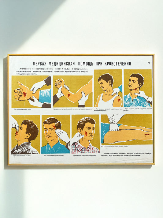 An original Soviet vintage framed educational medical instruction poster on a light wall, showing illustrated steps for first aid to a bleeding person in Russian, used at Civil Defence school classes in USSR. 